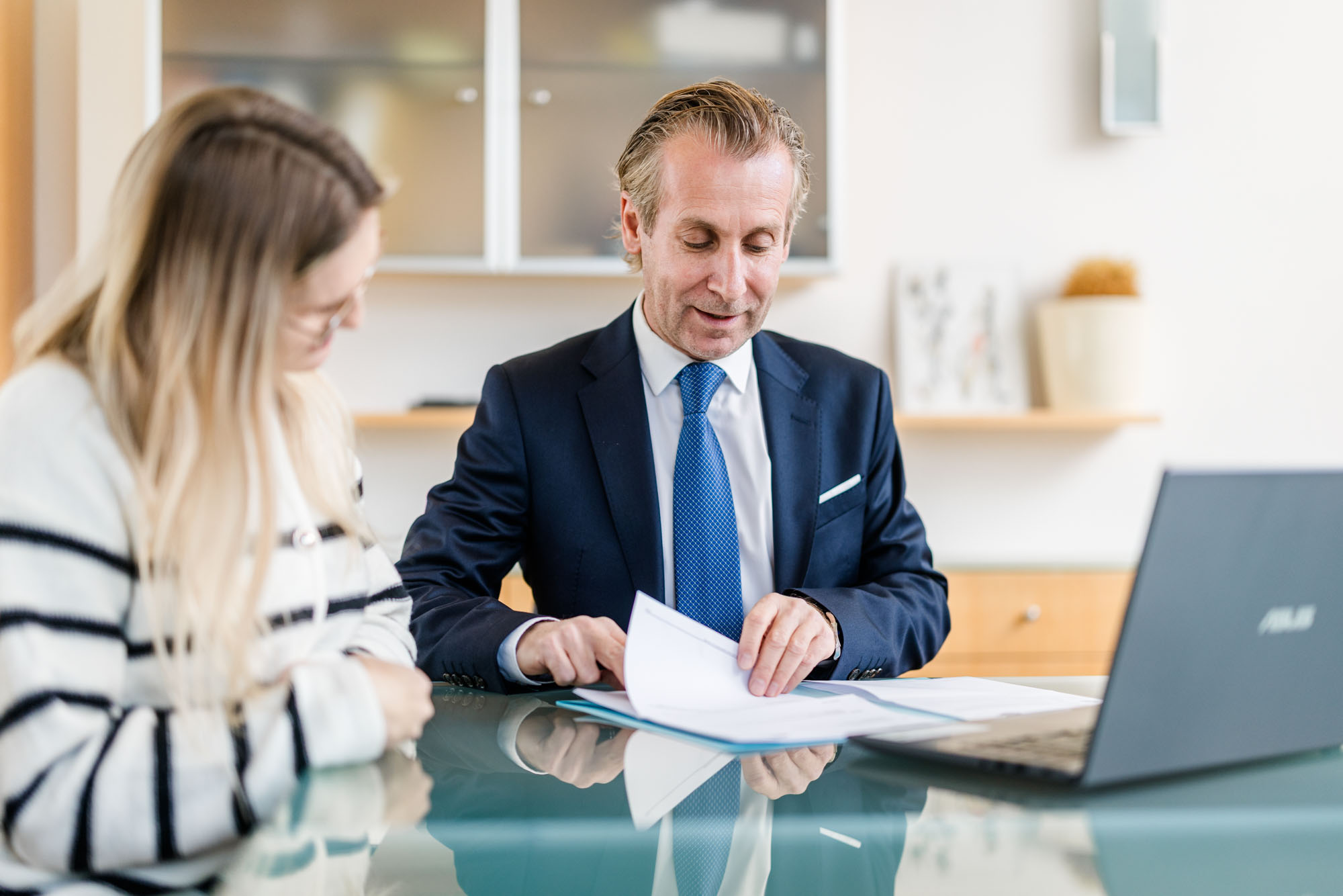Mann im Anzug hält Dokumente in der Hand und spricht mit einer Dame am Tisch